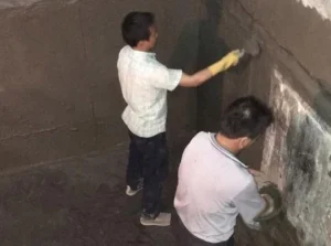 The picture shows two elevator shaft waterproofing workers applying waterproofing material to the elevator pit.